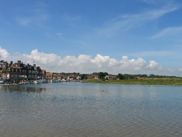 Blakeney at high tide, the harbour a broad sheet of water reaching the quay, with the village roofs and moored boats along the far side under a blue sky.