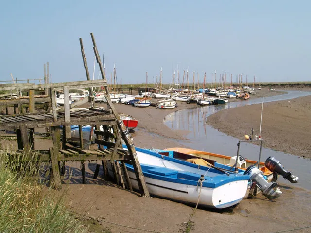 Blakeney at low water, small boats and dinghies standing on drying mud beside old timber staging, with a thin curve of remaining water threading away through the saltmarsh.
