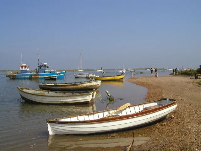 Brancaster Staithe with the tide in: rowing boats and fishing boats afloat on flat blue water right up to a shingle spit, people standing at the water’s edge in the sun.