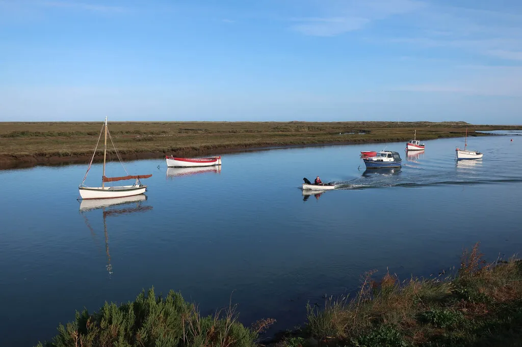 Overy Creek full of water under a clear sky, with a dinghy under outboard heading down the channel past moored boats lying at anchor, saltmarsh flat and green on either side.