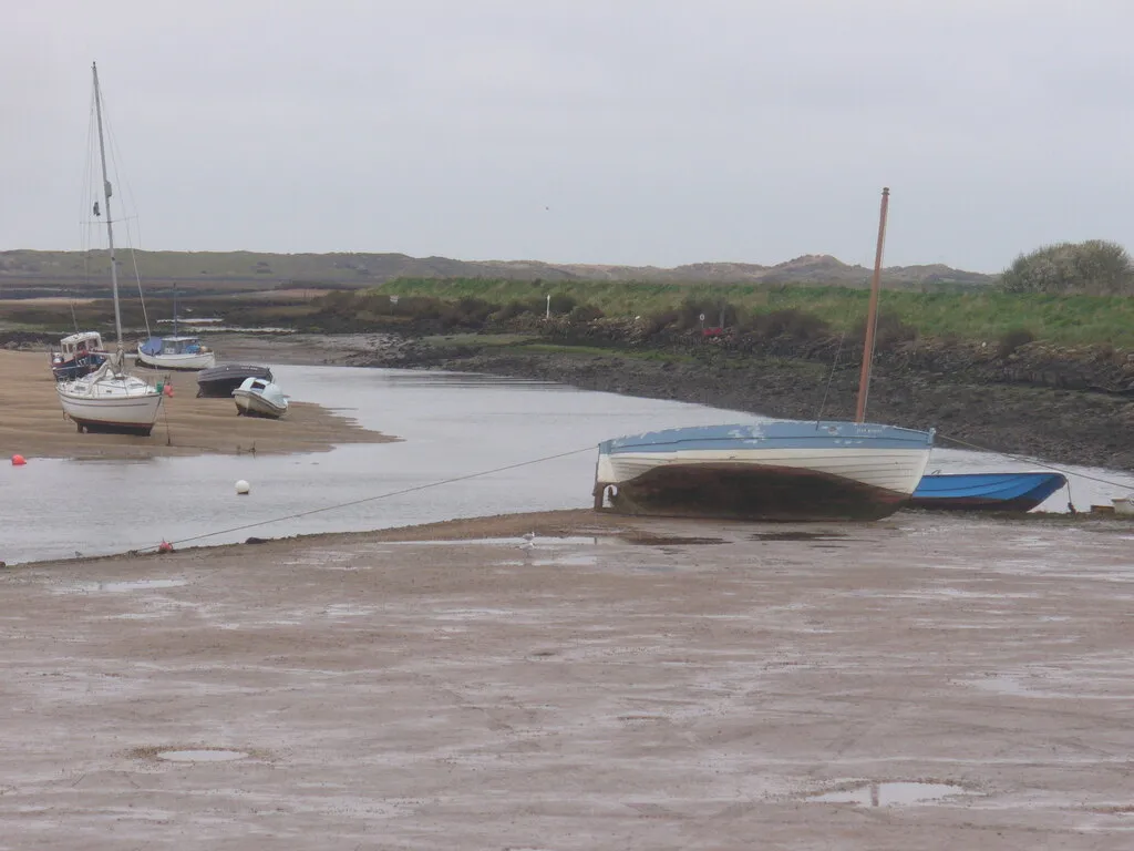 The same creek at low water on a grey day, reduced to a narrow ribbon between banks of mud, with a blue and white boat lying over on its bilge and yachts standing upright on the hard sand behind.