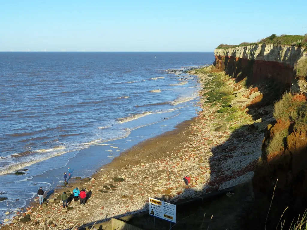 The banded red and white cliffs at Hunstanton with the sea right up against them, waves breaking along a narrow strip of shingle where a few people are walking.