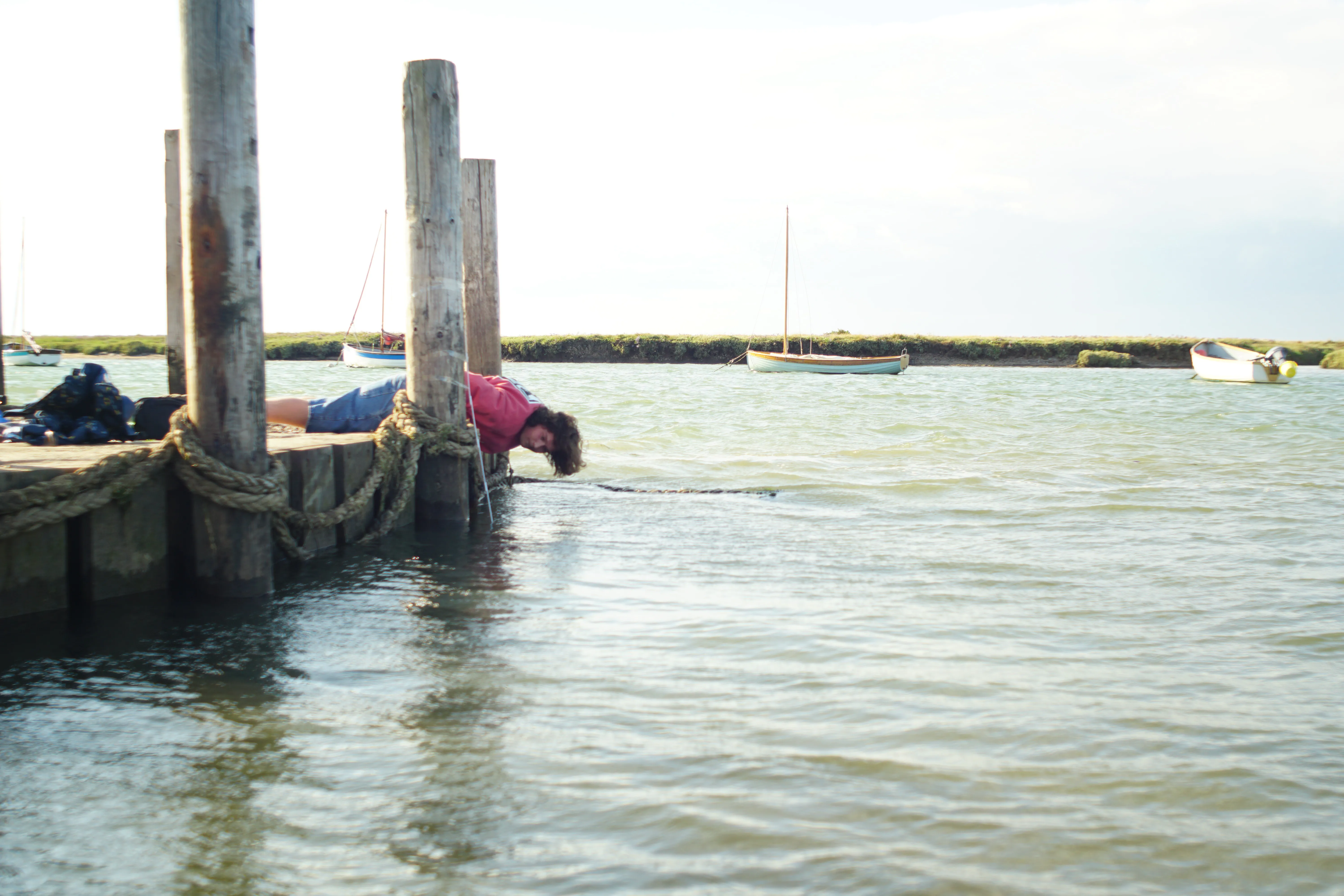 An observer lying flat on the quay at Burnham Overy Staithe with their head over the edge, reading the waterline against a measuring stick fixed to the timber, while moored boats sit afloat in the creek behind.