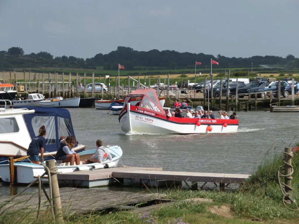 A loaded seal-trip boat under way down Morston Creek with passengers aboard in lifejackets, water high against the staging, cars parked along the quay behind.