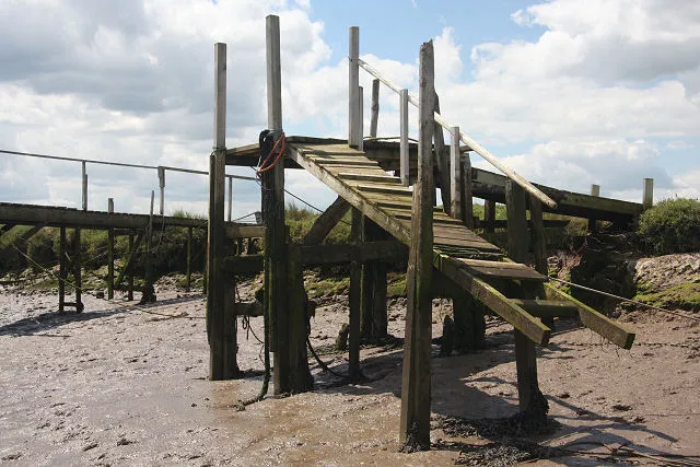 A weathered wooden landing stage at Morston standing on its legs above bare mud at low water, its steps and gangway ending over dried creek bed with no water beneath.