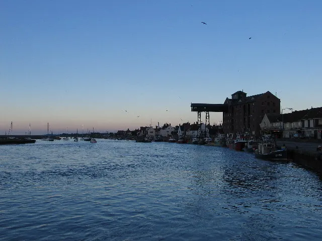 Wells harbour channel at dusk brimming with water, gulls over the surface, the quay and the tall granary with its gantry standing in silhouette against a pale sky.