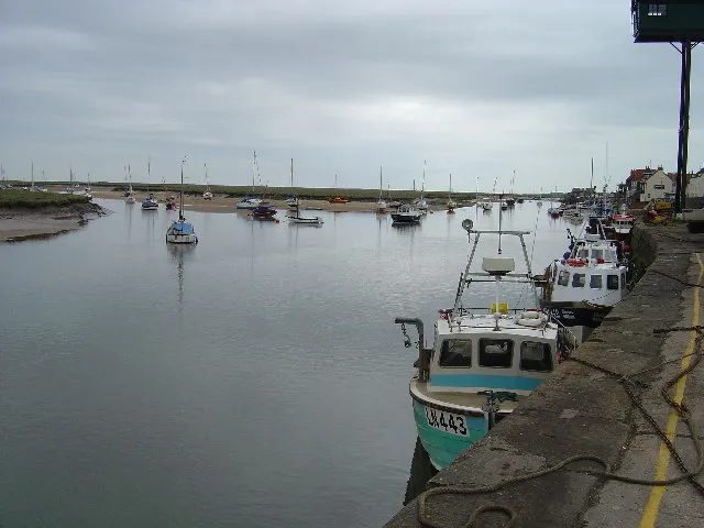 The same channel on an overcast day with the water withdrawn to a central strip, fishing boats tied along the quay wall sitting low, and moored craft beyond resting on exposed banks of mud.
